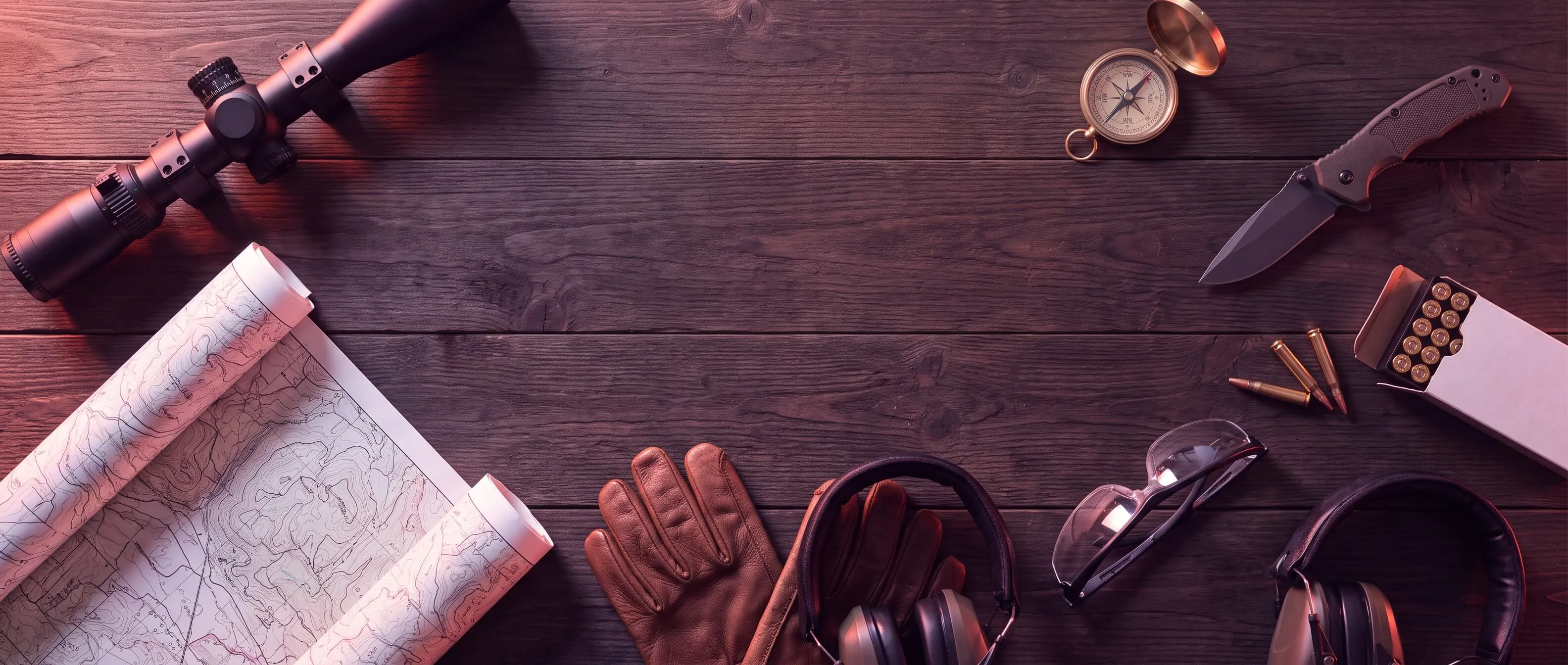 Rifle scope, map, compass, knife, gloves, ear protection, and ammunition laid out on a wooden table.