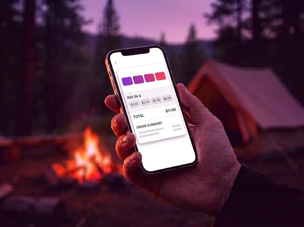 Hand holding a phone at a campsite showing a Pay in 4 checkout option.